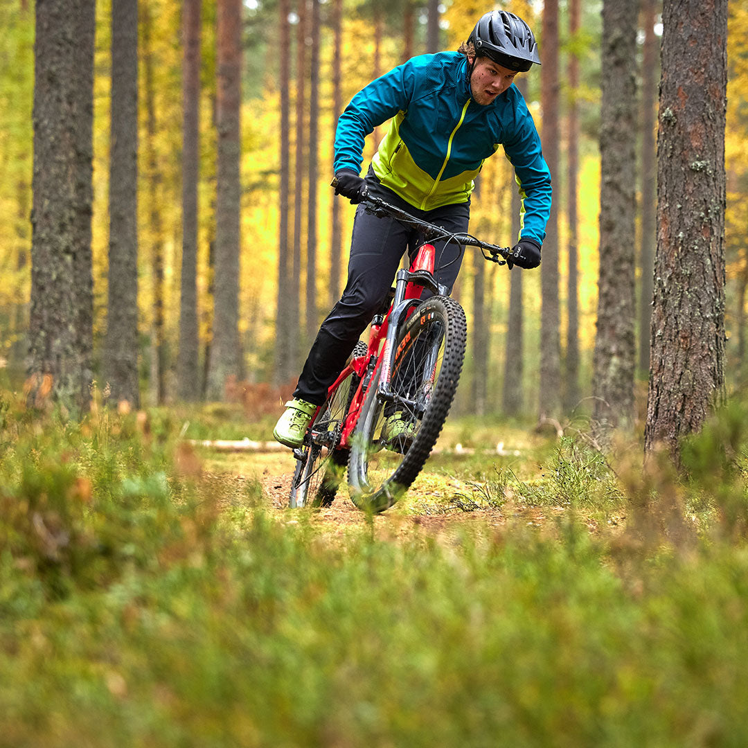 En person i en Halti Suunta Herr Hybrid-jacka och hjälm cyklar på en mountainbike på en skogsstig kantad av höga träd.
