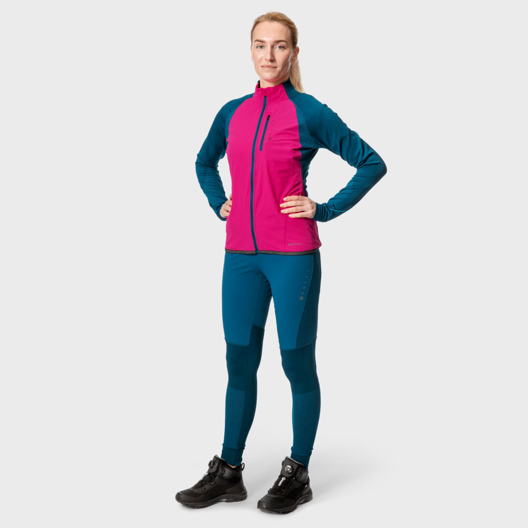 En person vänder sig framåt i Halti Tracks Seamless Tights Dam, rosa och blå, med svarta stövlar på en enfärgad ljus bakgrund.