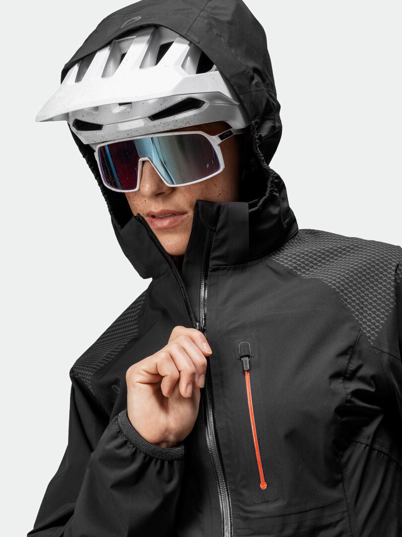 Personen bär en svart jacka med huva, en vit cykelhjälm och reflekterande solglasögon. Jackan har ett orange blixtlås på bröstet. Personen drar upp blixtlåset.