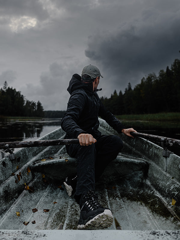 En person klädd i mörka friluftskläder och keps ror en metallbåt på en lugn sjö, omgiven av träd under en molnig, mulen himmel.