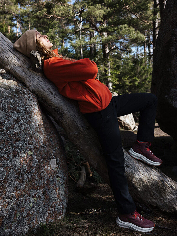 En person i röd jacka och solbrun mössa, klädd i Halti Rapid DX Mid Outdoorskor Dam, lutar sig mot ett nedfallet träd i skogen.
