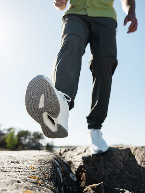 En person klädd i Halti Syke Sneaker Herr och mörka byxor går fram på stenar under en klar himmel.