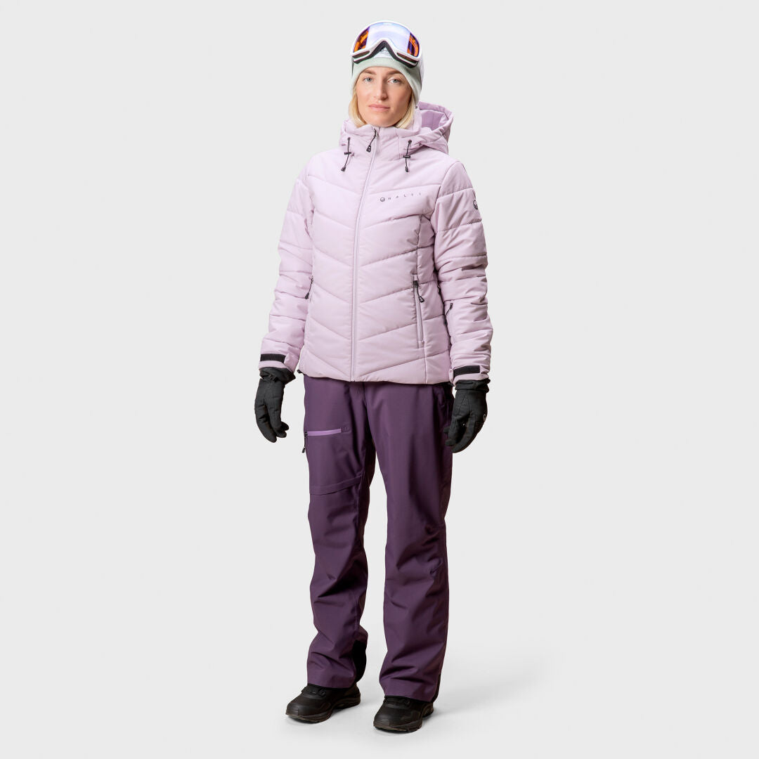 En person bär en ljuslila Halti Mellow Puffer Skidjacka Dam, mörklila snöbyxor, handskar och skidglasögon.