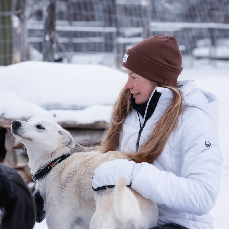 Iklädd en Halti Mellow Puffer Skidjacka Dam och brun mössa klappar en person en vit hund på en snöig, inhägnad gård.