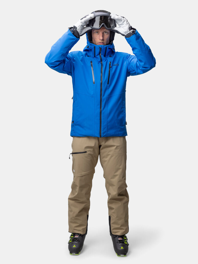 Man i Halti Vertica DX Skidjacka och solbruna byxor justerar skidglasögon, bär handskar och pjäxor, vänd framåt.