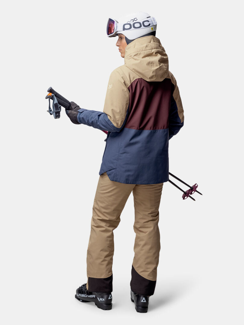 En person i en Halti Gracer DX Skidjacka Dam står med skidstavar i handen och vänder sig bort mot en enfärgad bakgrund.