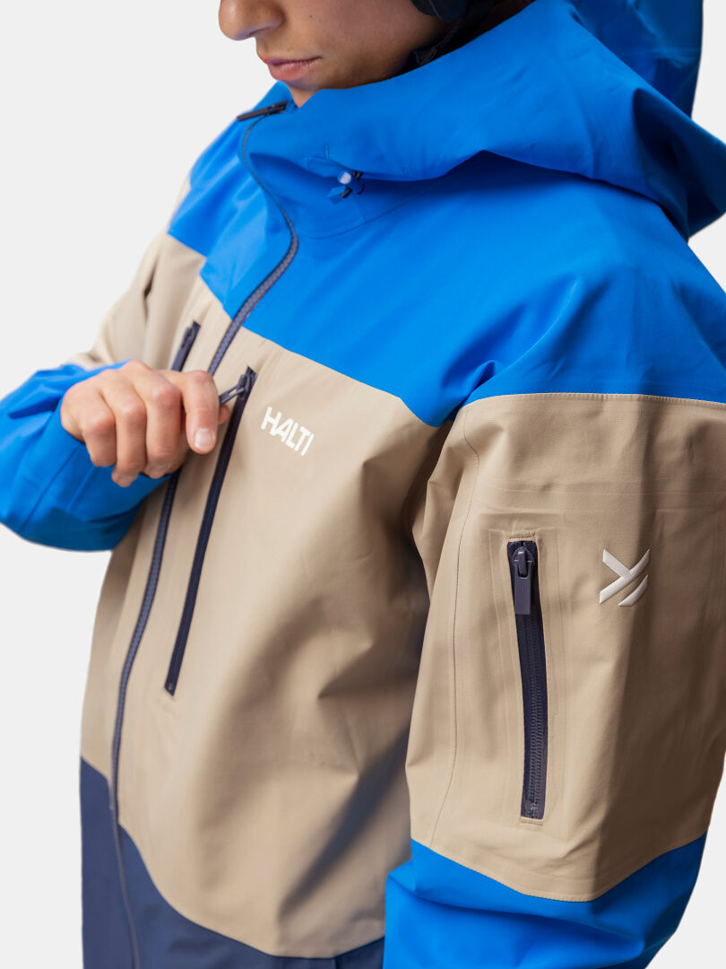 En person drar upp dragkedjan i en brunblå Halti Rizle 3L DX Skidjacka Unisex-jacka med huva; en ficka med dragkedja i ärmen syns.