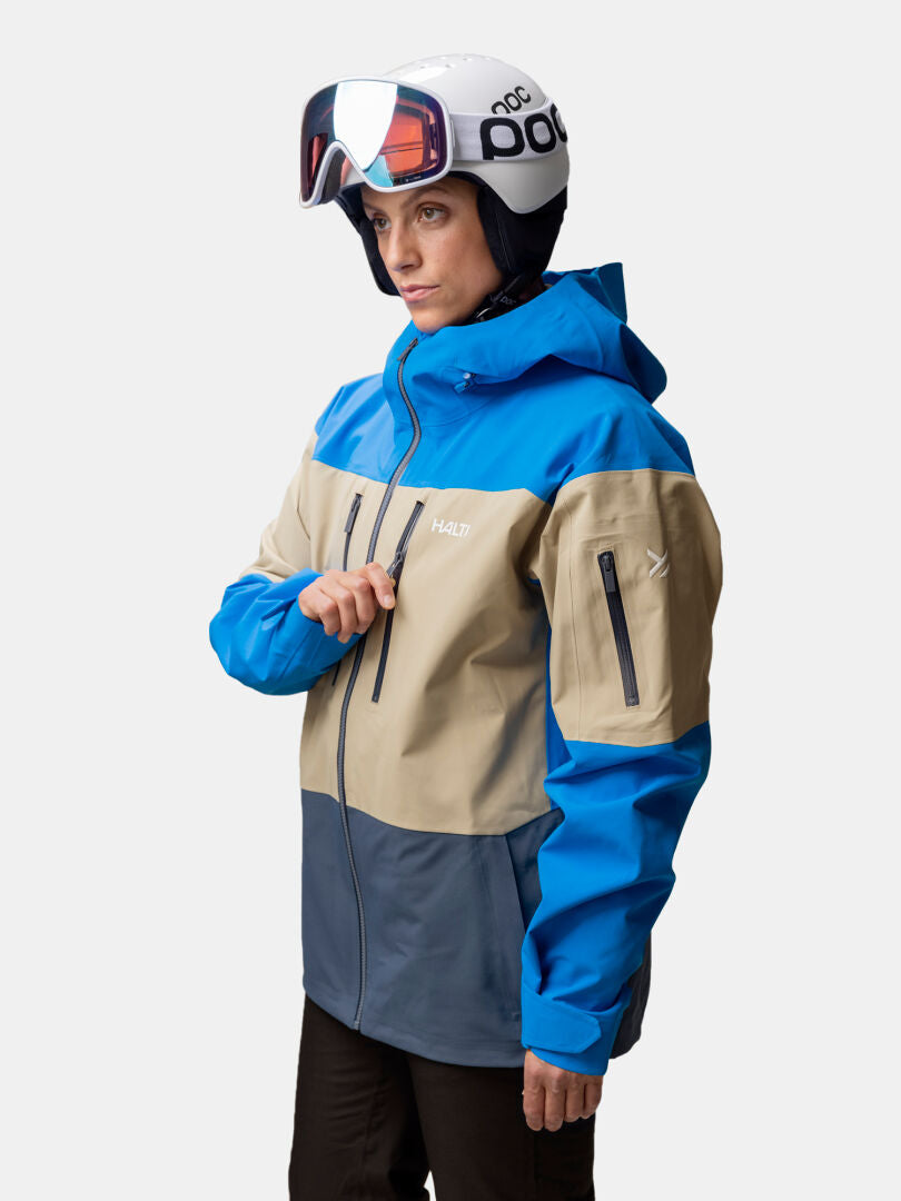 En person i blå och beige Halti Rizle 3L DX Skidjacka Unisex och hjälm står till vänster och håller i dragkedjan.