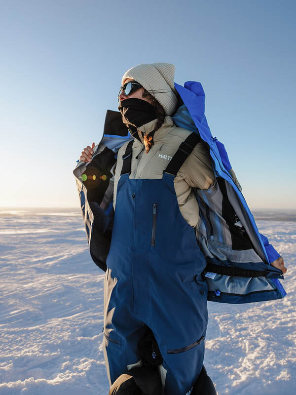 En person som bär en Halti Rizle Unisex 3L DX Skidjacka över ett beige, isolerat lager och blåa hängselbyxor i ett snötäckt bergslandskap.