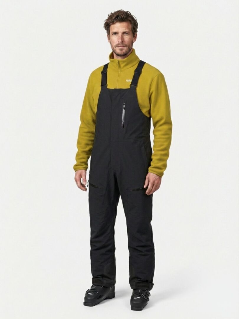 En man klädd i svarta Rizle 3L DX Skidbyxor Unisex-skidbyxor med hängslen över en gul fleecetröja och skidkängor.