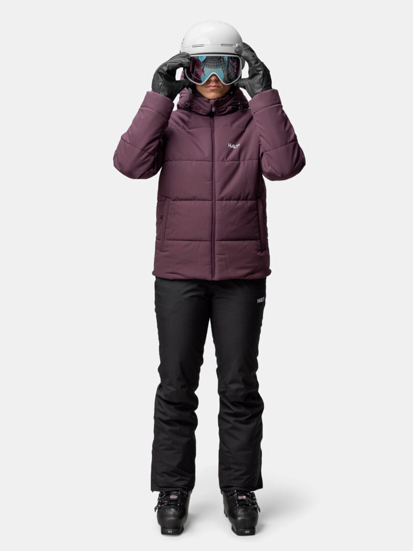 En person bär Halti Ascale Puffer Skidjacka Dam, svarta snöbyxor, pjäxor, handskar, hjälm och skyddsglasögon vända framåt.