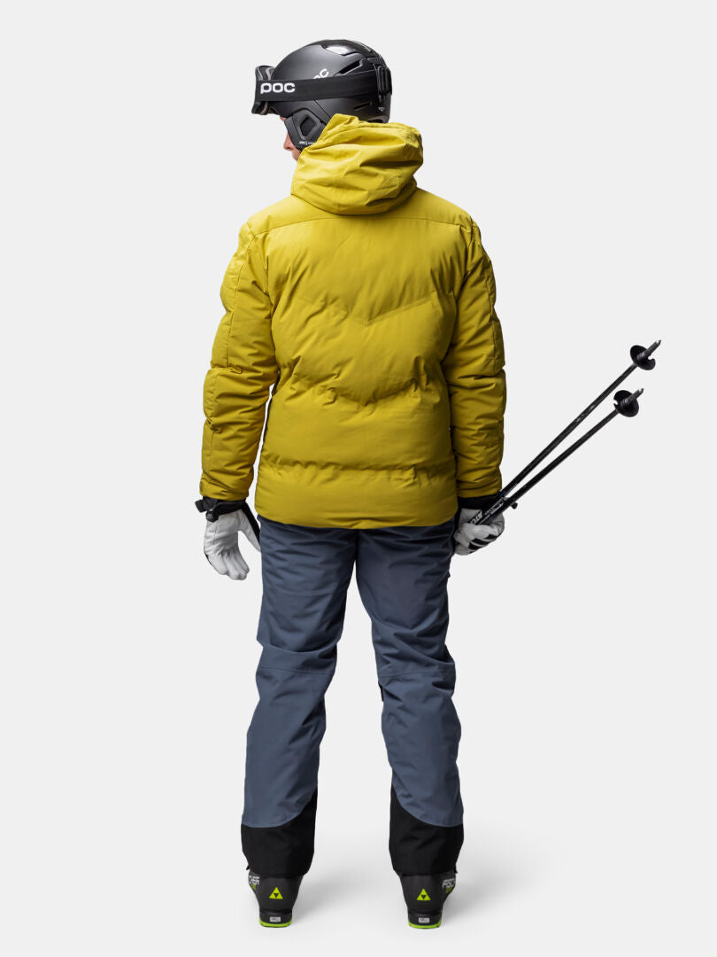 Person i Halti Nordic Arcty II Skijacka Herr, blå byxor och skidutrustning står vänd bort och håller i skidstavar.