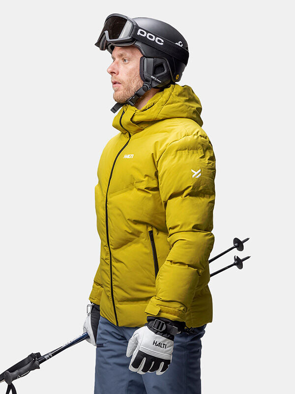 En man bär en Halti Nordic Arcty II Skijacka Herr, hjälm och skyddsglasögon och håller i skidstavar mot en vit bakgrund.