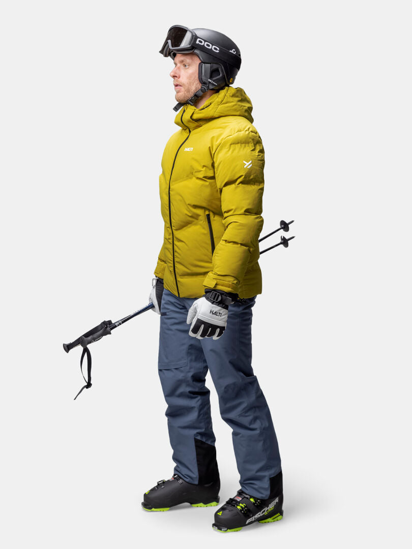 En person i gul Halti Nordic Arcty II Skijacka Herr, blå byxor, pjäxor, hjälm, skidglasögon och skidstavar.