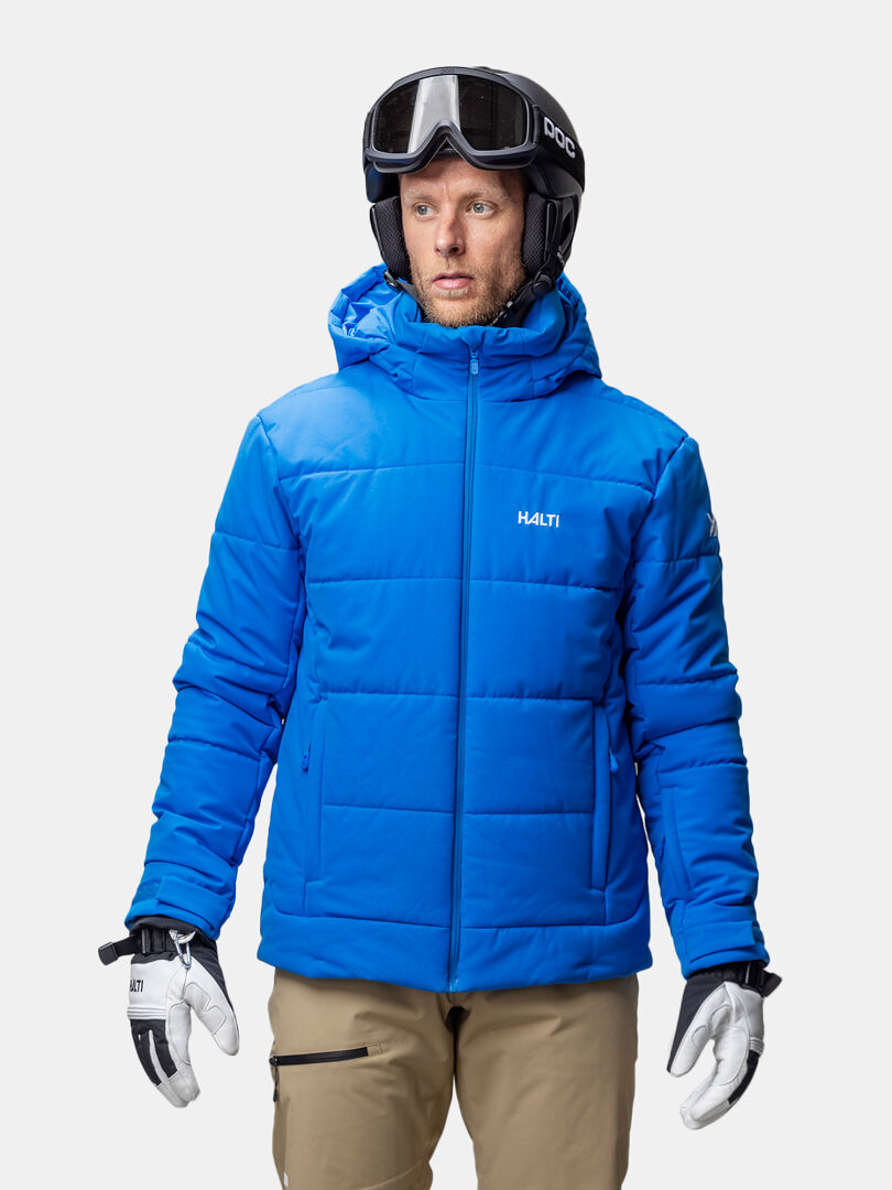 En man i Halti Ascale Puffer Skidjacka Herr, solbruna byxor, skidhandskar och skidglasögon står mot en vit bakgrund.