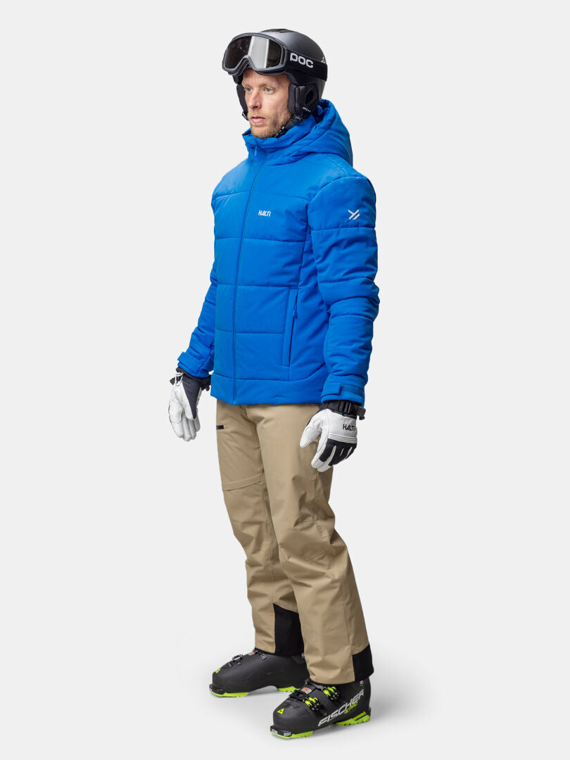 En person i blå Halti Ascale Puffer Skidjacka Herr, beige byxor, pjäxor och hjälm står vänd åt höger.