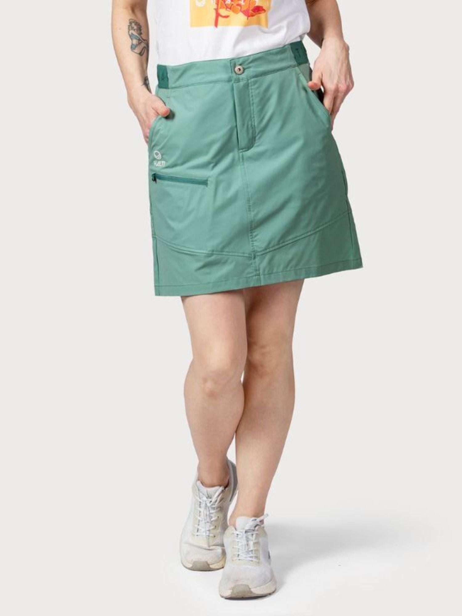 Person som bär en grön Halti Pallas Dam Lite X-stretch Kjol, vit t-shirt med grafik och vita sneakers med händerna i fickorna.