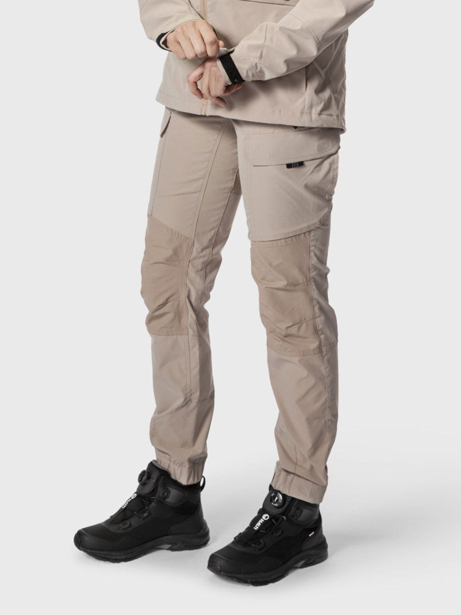 En person i Halti Hiker Dam Lite Outdoor Byxor och en beige jacka i återvunnen nylon står och har svarta vandringsskor på sig.