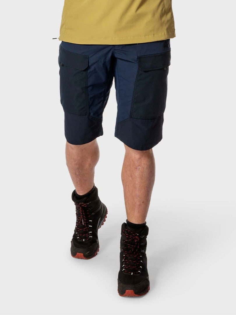 Personen bär Halti Hiker Herr Lite Shorts i marinblå färg, gul skjorta och svarta kängor mot en ljus bakgrund.