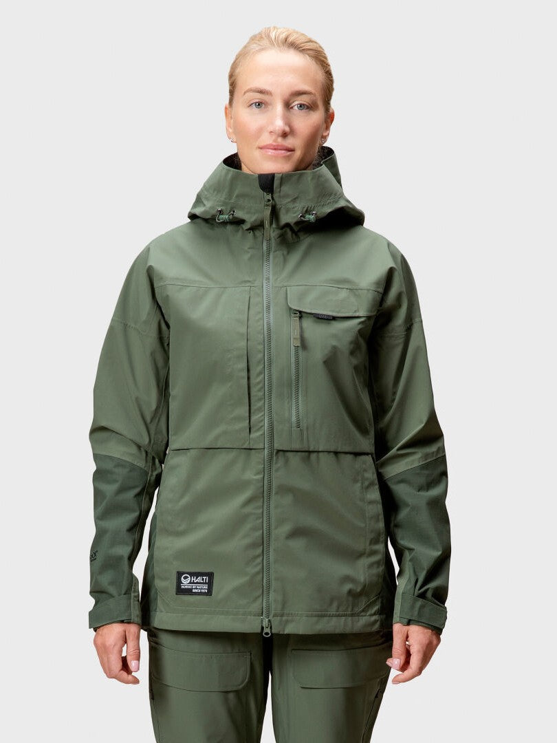 En person i grön Halti Hiker DrymaxX 3L Ventilated Jacka Dam och matchande byxor står mot en ljus bakgrund.