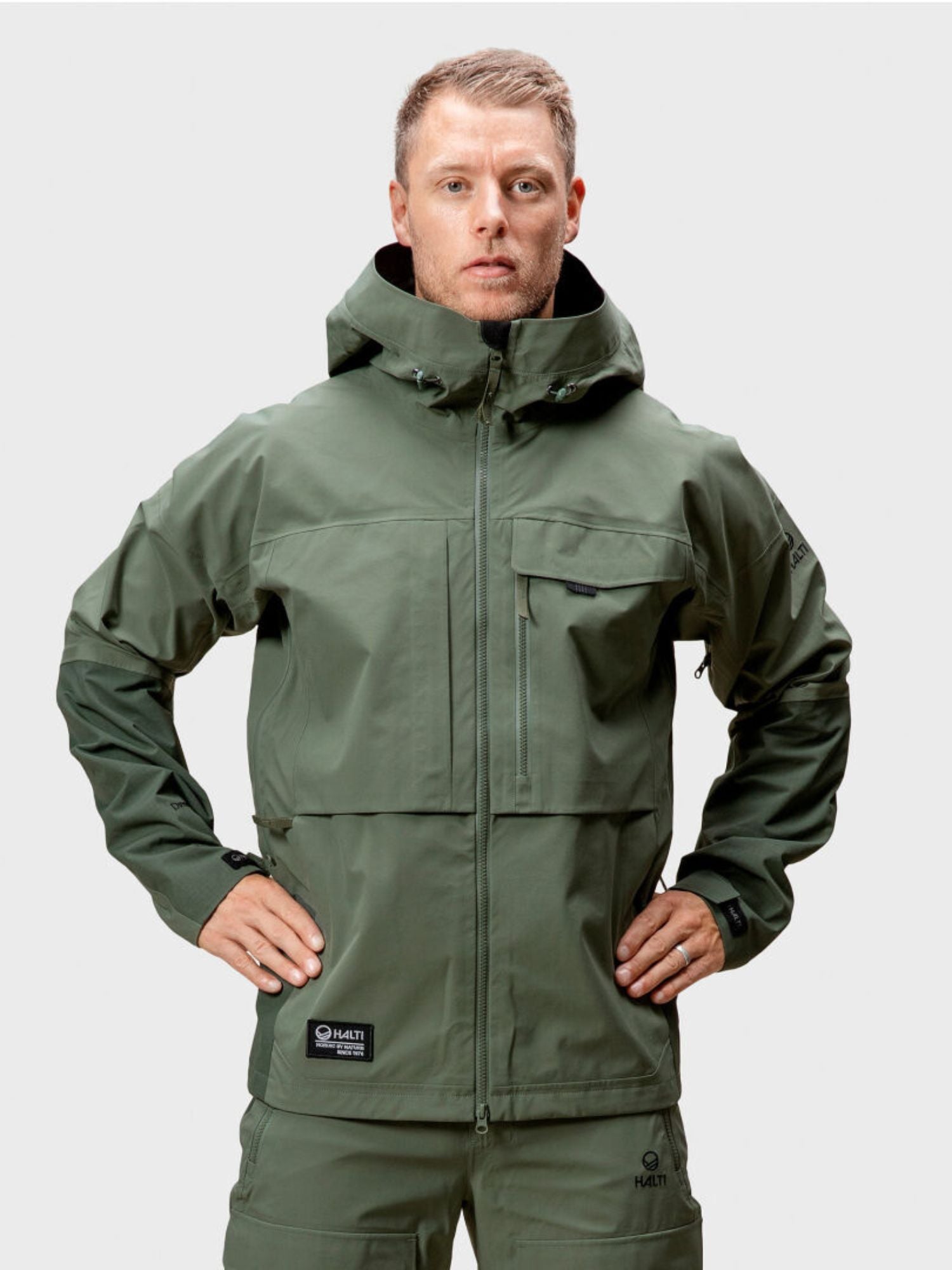 En man i grön Halti Hiker DrymaxX 3L Ventilated Jacka och matchande byxor står vänd framåt med händerna på höfterna.