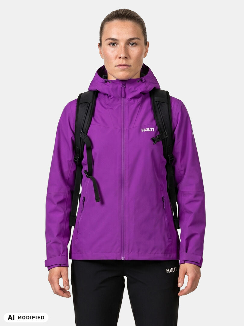 En person i en Halti Forter DX Skaljacka för damer, lila, med en svart ryggsäck, stående mot en ljus, enfärgad bakgrund.