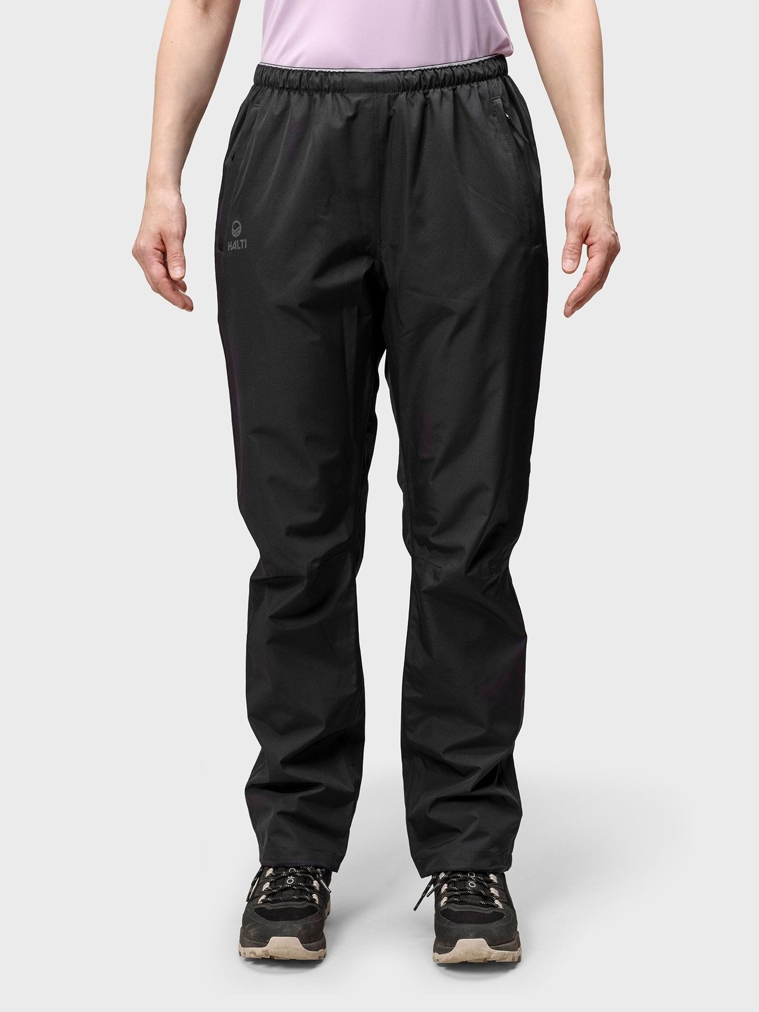 En person i svart Halti Forter DX Skalbyxor Dam, svarta sneakers och en ljuslila skjorta står mot en enfärgad bakgrund.