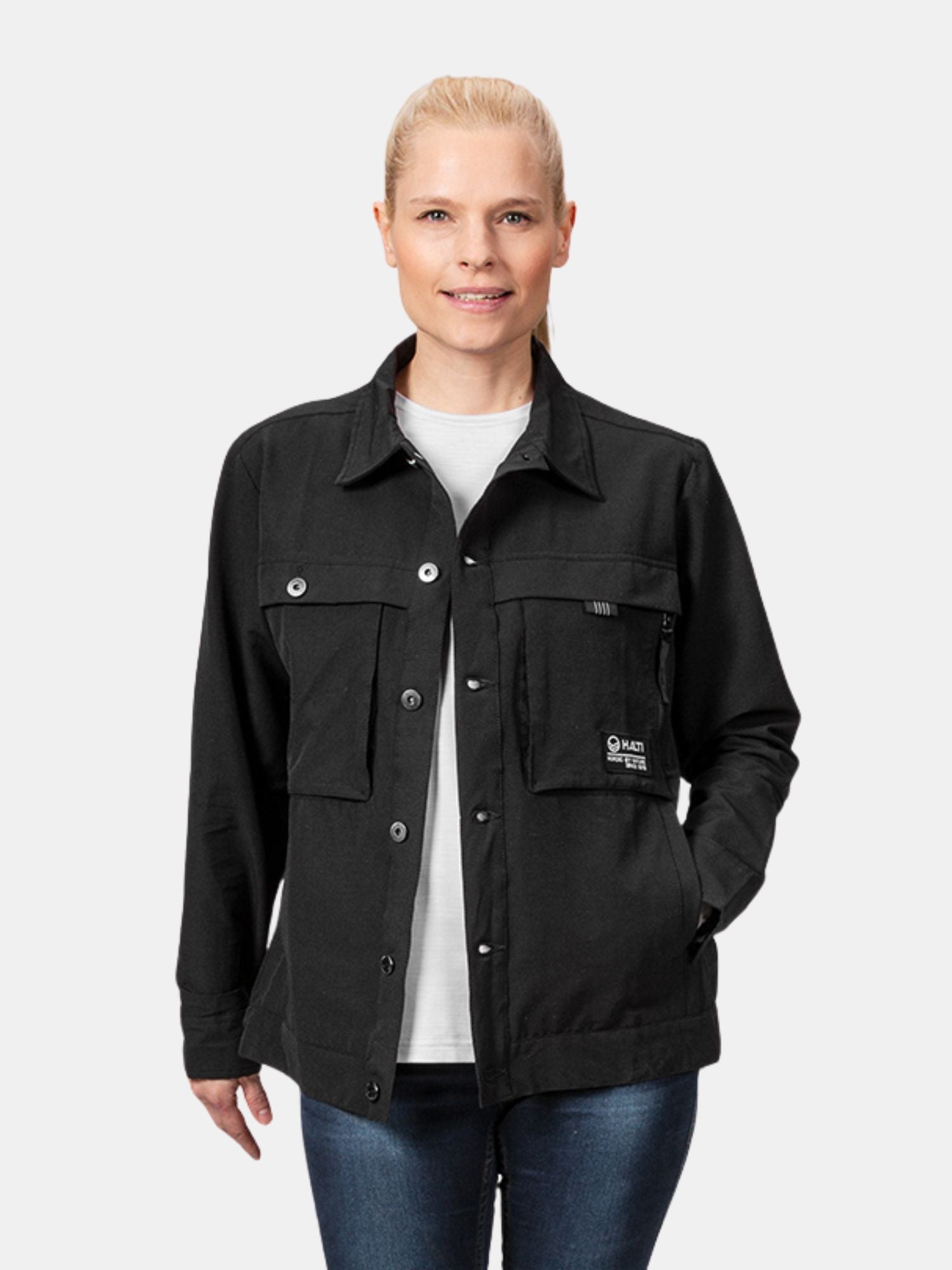 En kvinna i en Halti Easy Hike Skjortjacka Dam med flexibelt tyg och blå jeans står mot en enfärgad ljus bakgrund.