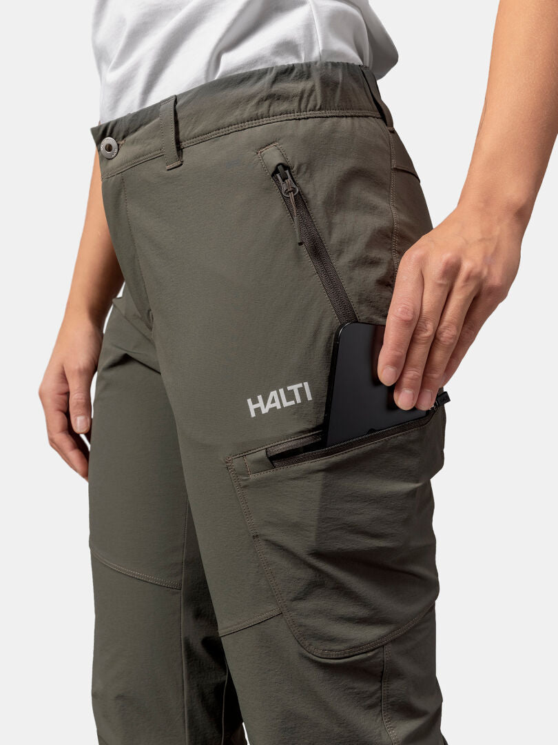 Person som lägger en svart smartphone i den dragkedjeförsedda sidofickan på olivgröna Halti Pallas Evo X-stretch Byxor Dam.