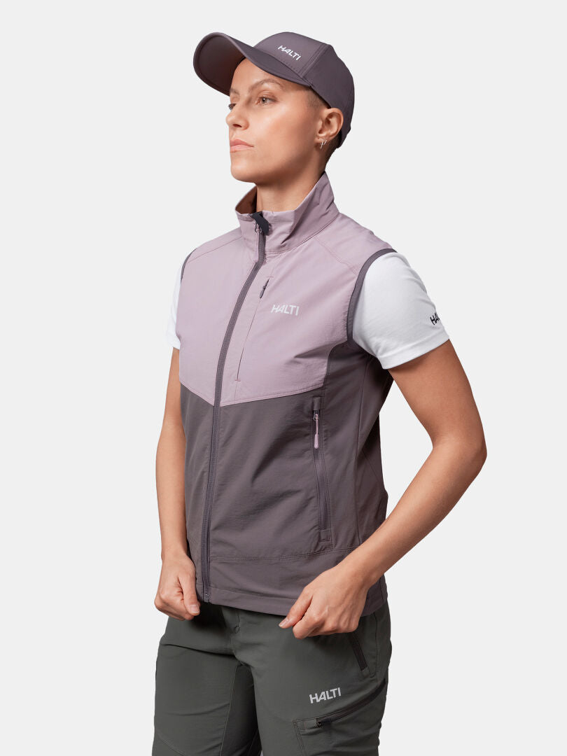 Kvinna i Halti Pallas Evo X-stretch Väst, vit t-shirt, grå keps och mörka byxor står i sidled med händerna på höfterna.