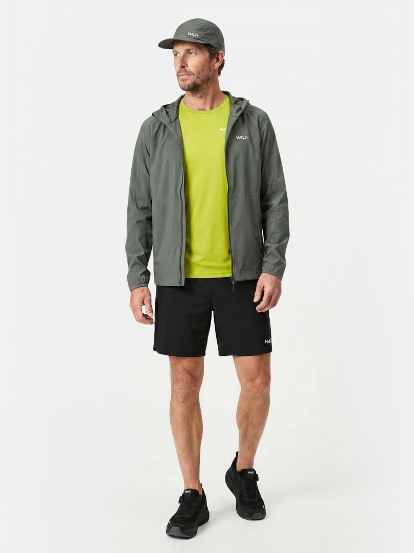 En man i grå Halti Pisarat 2,5L DX Skaljacka Herr, limegrön skjorta, svarta shorts och keps står vid en enfärgad bakgrund.