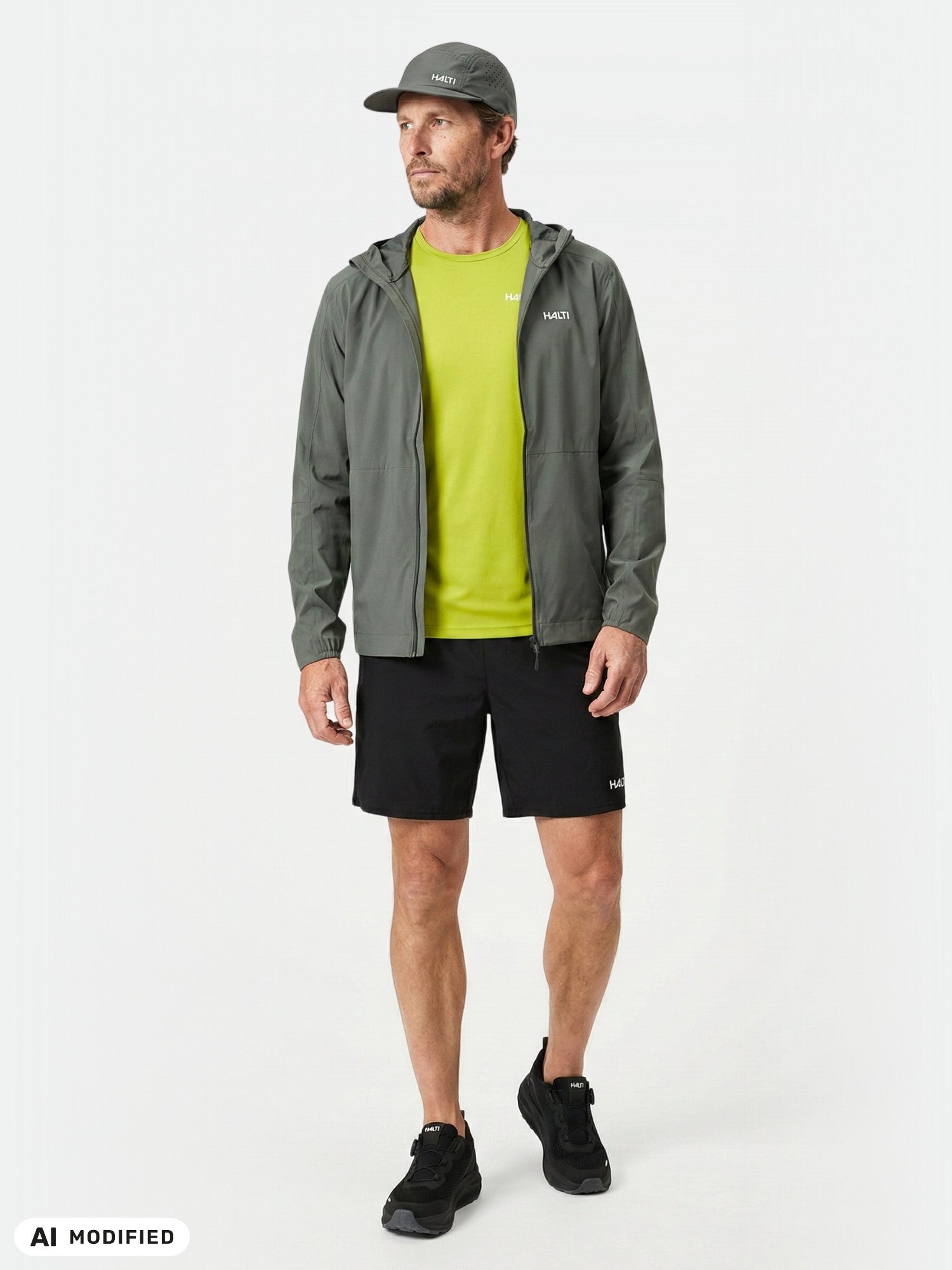 Mannen bär en Halti Pisarat 2,5L DX Skaljacka Herr i grått, en limegrön skjorta, svarta shorts och skor samt en grå keps.