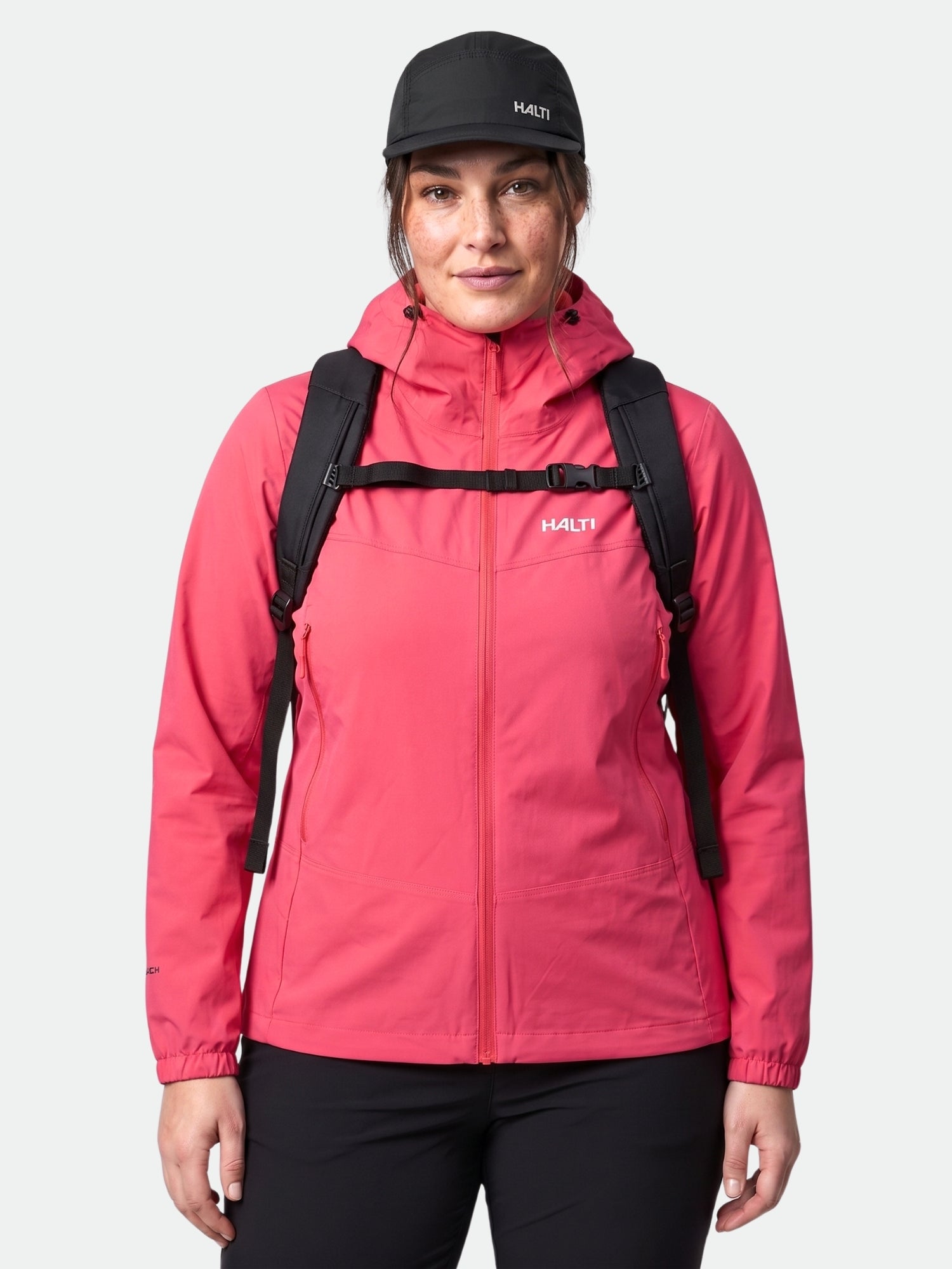 En person klädd i en röd Halti Pallas Evo X-stretch Jacka Plus Dam, en svart ryggsäck och en keps vänder sig mot kameran.