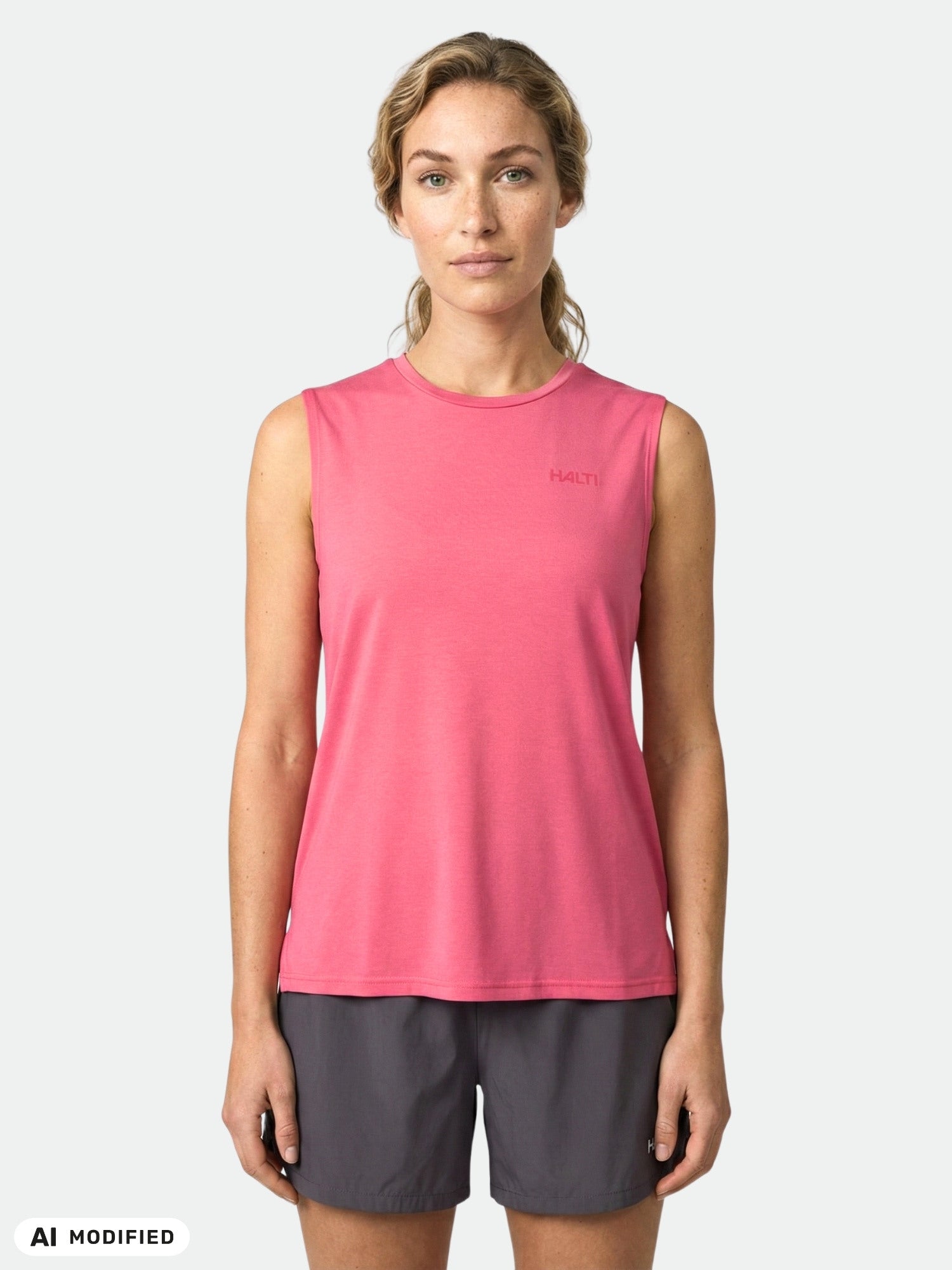 En kvinna står vänd framåt i en rosa Halti Kalteva Top Dam och mörka shorts mot en enfärgad, ljus bakgrund.