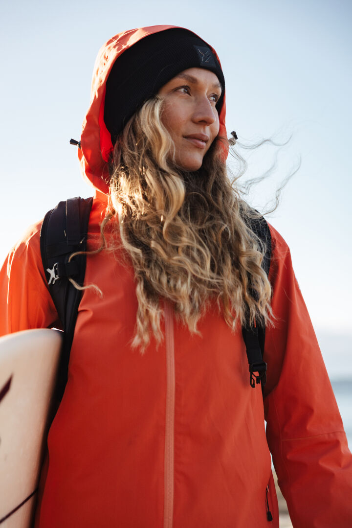 Kvinna i orange Staala DX Skaljacka Dam från Halti och svart beanie håller en surfbräda, blickar mot himlen.
