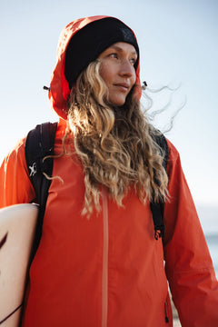 Kvinna i orange Staala DX Skaljacka Dam från Halti och svart beanie håller en surfbräda, blickar mot himlen.