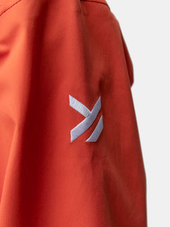 Närbild av Halti Staala DX Skaljacka Dam i orange med en vit broderad geometrisk logotyp med tre vinklade linjer.