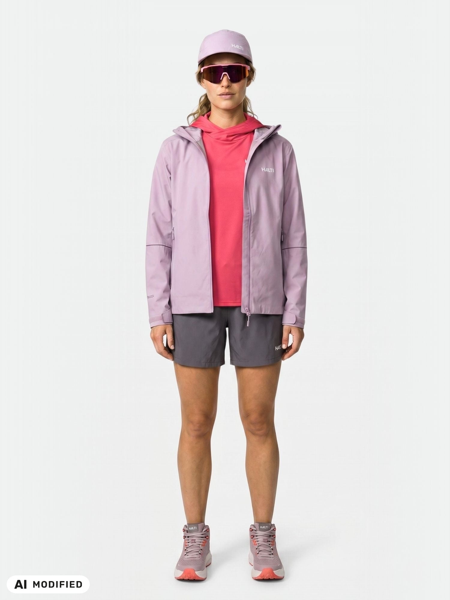 Modellen bär en Halti Staala DX Skaljacka för damer, en röd tröja, grå shorts, en rosa keps och sneakers och står med blicken riktad framåt.