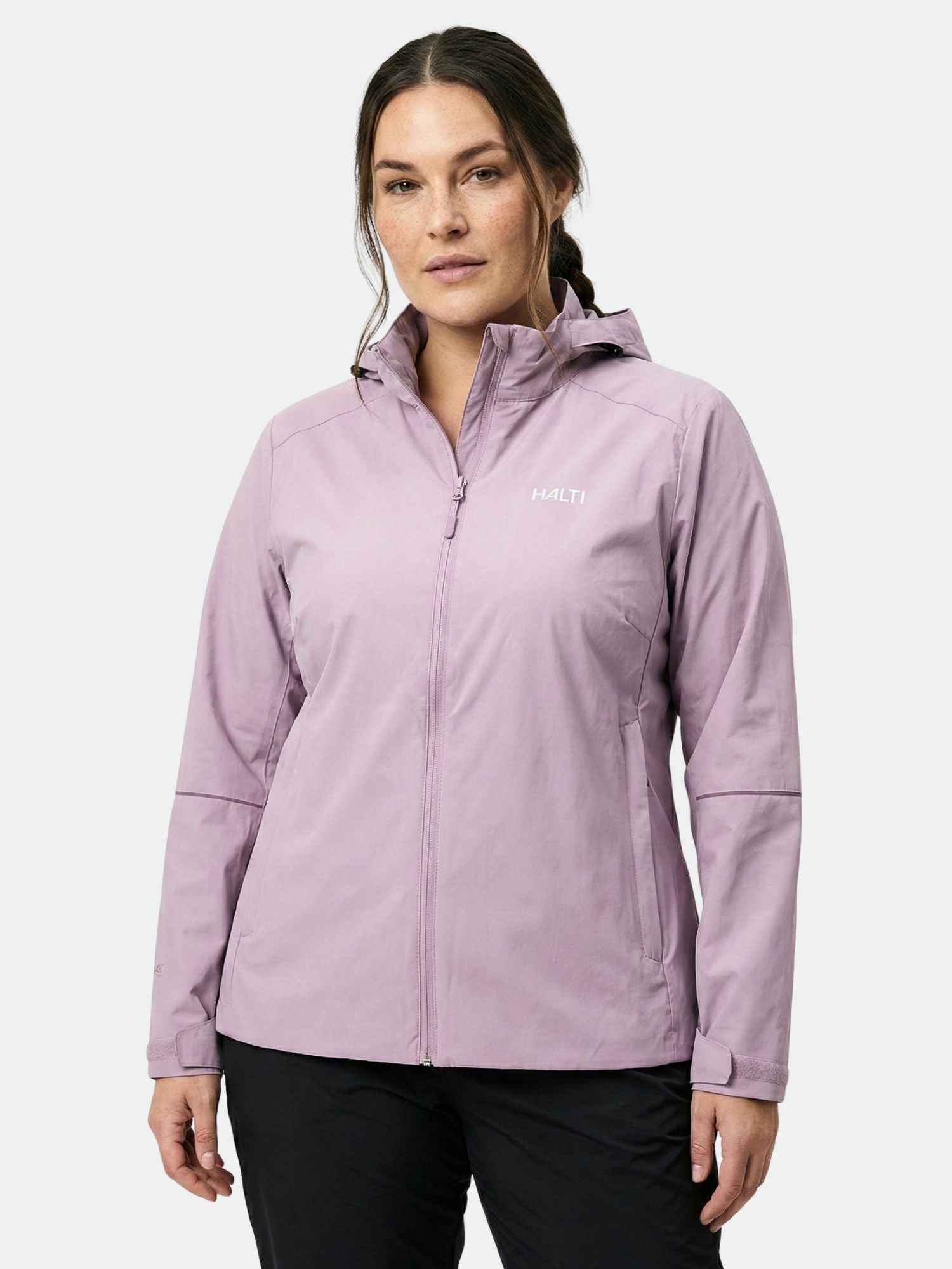 En kvinna bär en ljuslila Halti Staala DX Skaljacka Dam Plus med huva och står mot en ljus bakgrund.