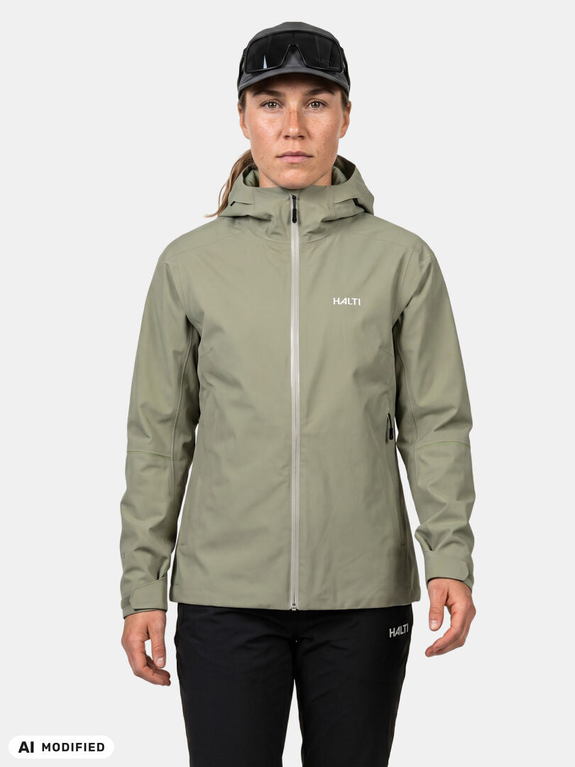 En person klädd i en ljusgrön Halti Staala DX Skaljacka Dam och svarta byxor, stående mot en enfärgad bakgrund.