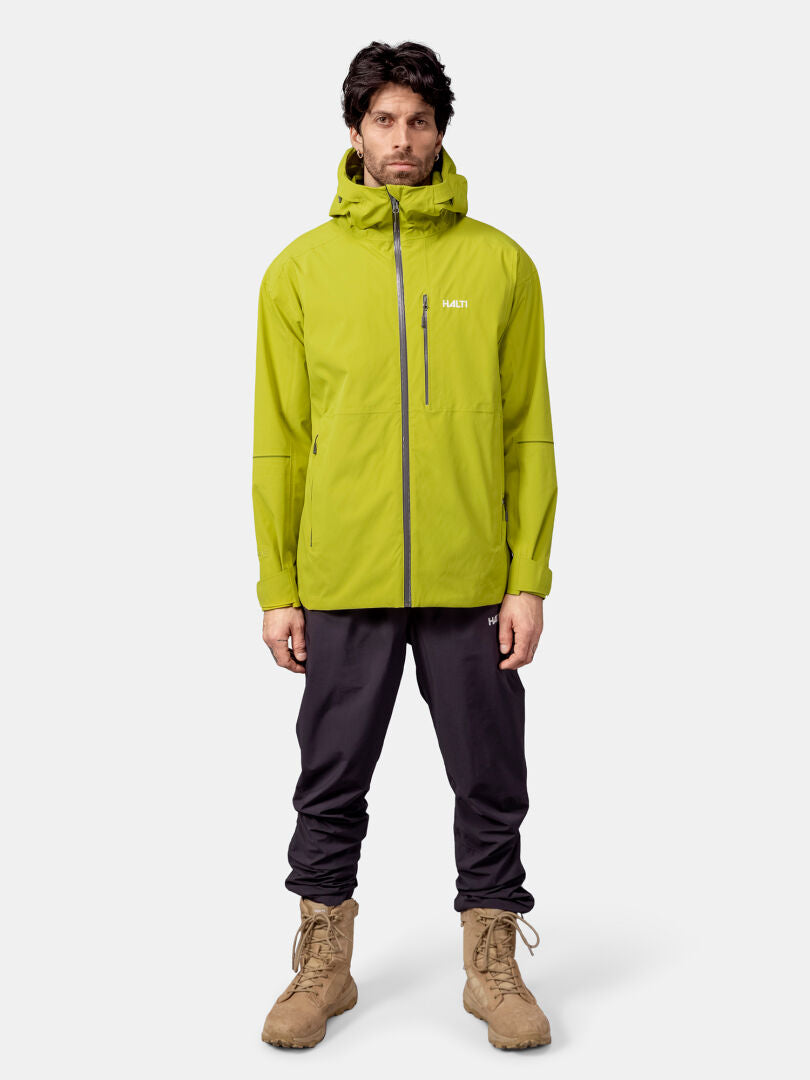En man tittar fram och har på sig en Halti Staala DX Skaljacka Herr i ljusgrönt, svarta byxor och bruna snörkängor.