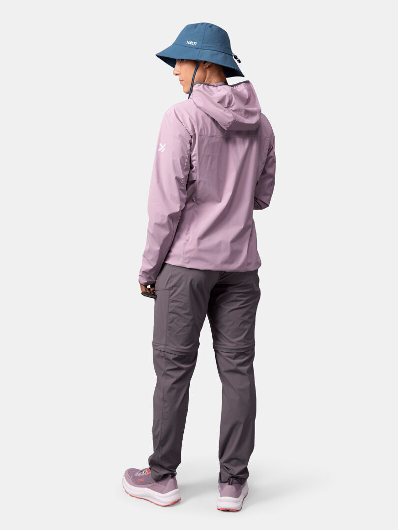 En person i blå mössa, Halti Pallas Cool X-stretch Jacka Dam, grå byxor och sneakers står med ansiktet vänt bort.