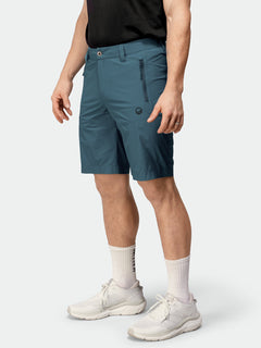 En person i Halti Pallas Cool X-stretch Shorts, svart skjorta, vita strumpor och sneakers står mot en enfärgad bakgrund.