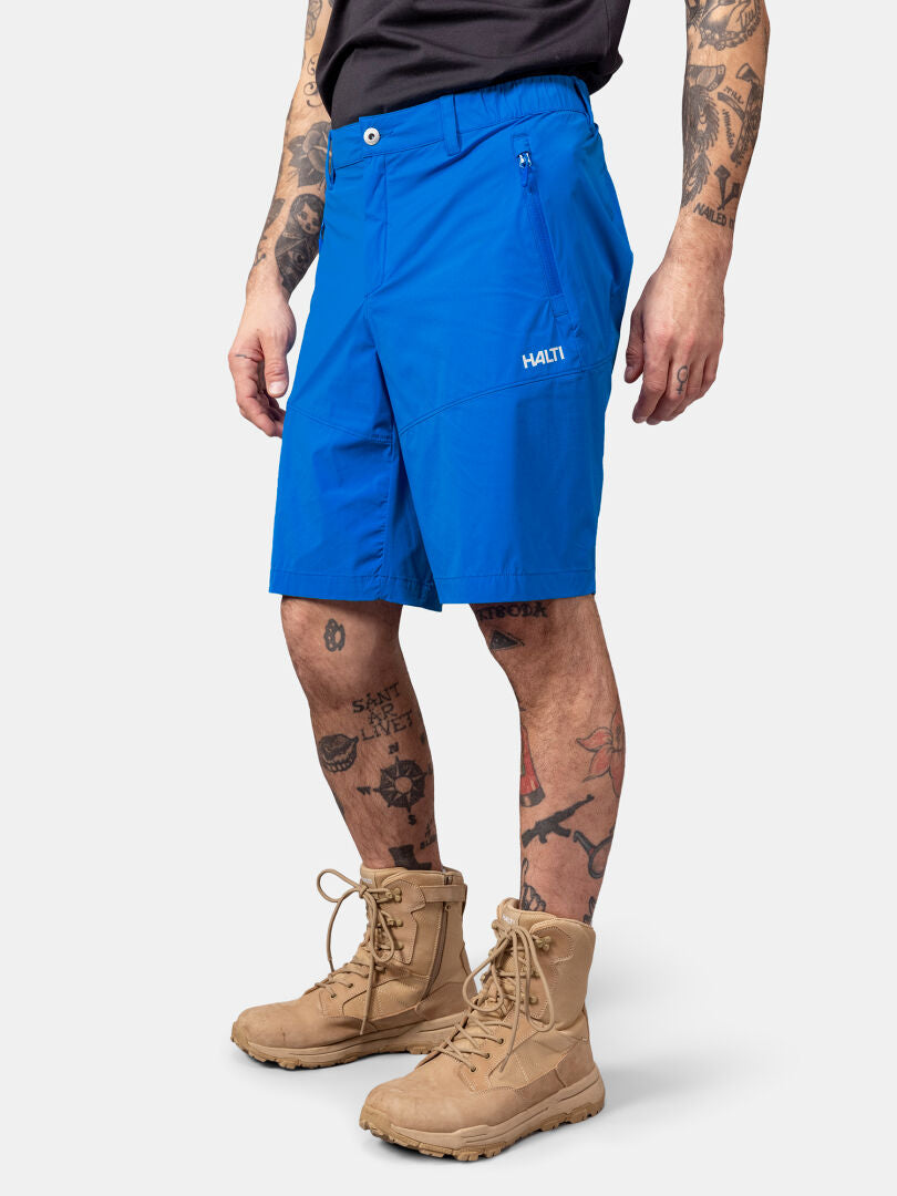 Person klädd i blå Halti Pallas Cool X-stretch shorts, bruna snörkängor, svart skjorta, med tatueringar på armar och ben.