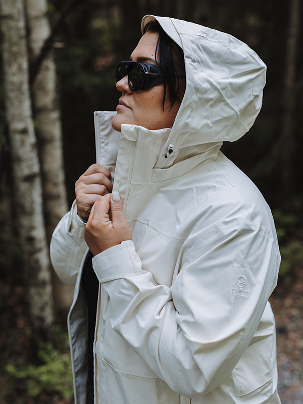 Kvinna i Halti Kallas DX Parka Plus Dam och solglasögon justerar kragen utomhus nära träd.