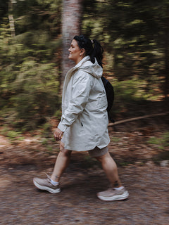 En person i en Halti Kallas DX Parka Plus Dam och sneakers går på en skogsstig, omgiven av träd och grönska.