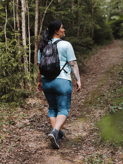 Person i Halti Pallas Cool X-stretch Capribyxor Dam Plus och ryggsäck promenerar längs en skogsstig bland träd och grönska.