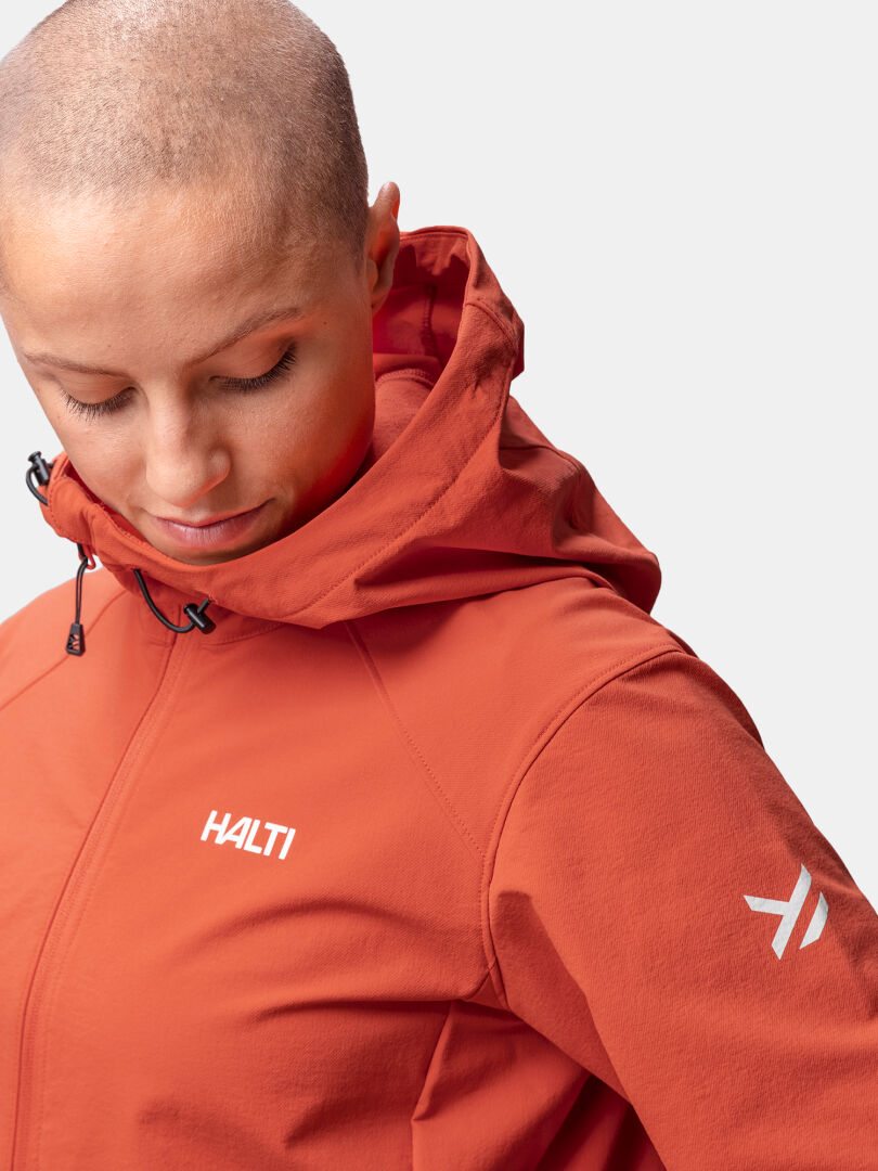 Person med rakat huvud bär en orange Halti Finholma X-stretch Jacka Dam och tittar ner mot en enfärgad bakgrund.