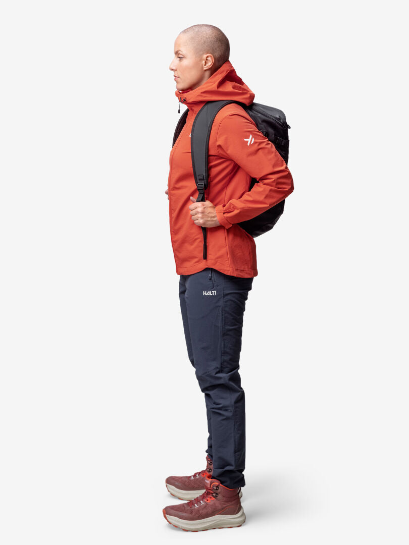 Person i Halti Finholma X-stretch Jacka Dam, mörka byxor, stövlar och svart ryggsäck står i profil på vit bakgrund.