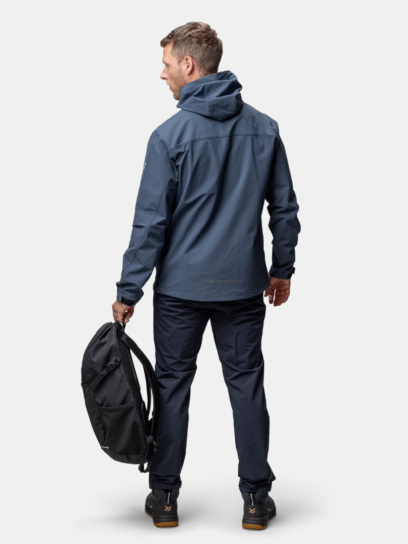 Man klädd i Halti Finholma X-stretch Jacka Herr i blått, mörka byxor och med en svart ryggsäck i handen - perfekt för utomhusäventyr.